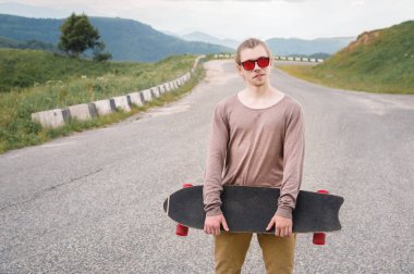 Şık bir genç adam gün batımından sonra akşam elinde bir paten veya longboard ile dolambaçlı bir dağ yolu boyunca ayakta. Gençlik spor ve seyahat hobileri kavramı
