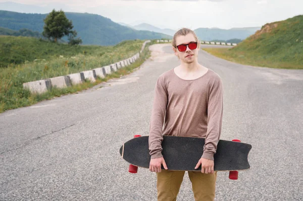 Ein stylischer junger Mann, der am Abend nach Sonnenuntergang mit Skate oder Longboard in der Hand an einer kurvenreichen Bergstraße steht. das Konzept des Jugendsports und der Reiselust — Stockfoto