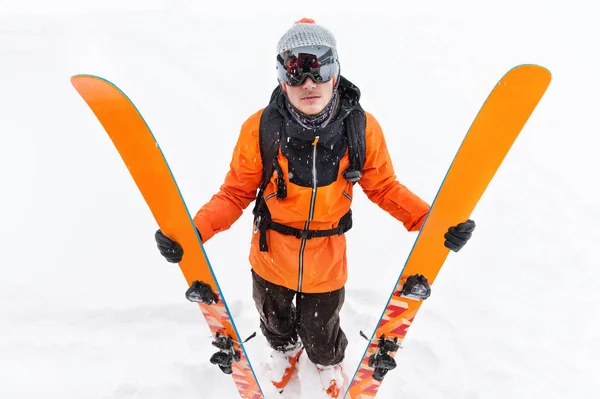 Профессиональный лыжник в оранжевом черном костюме с черной лыжной маской с лыжами в руках стоит с недовольной массой во время метели на светлом фоне в снегу — стоковое фото