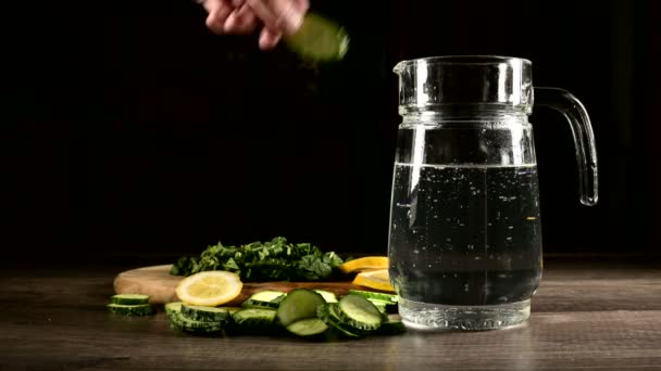 Sliced lemon cucumbers and mint leaves on a wooden cutting board next to a glass carafe with sparkling water. Male hand throwing cucumber slices into a glass of soda — Stock Video