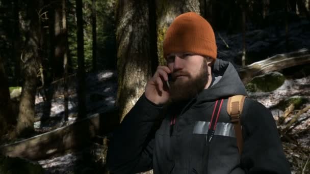 Ein bärtiger Mann mit Hut, der in einem Nadelwald mit einem Handy telefoniert. das Konzept der zellularen Kommunikation an schwer zugänglichen Orten — Stockvideo