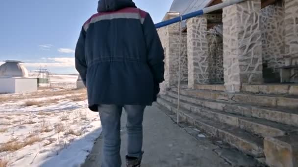 Seguindo um pesquisador masculino em jeans e um uniforme quente caminhando pela neve em um dia ensolarado de inverno entre os edifícios científicos do observatório solar. Estação de investigação científica — Vídeo de Stock