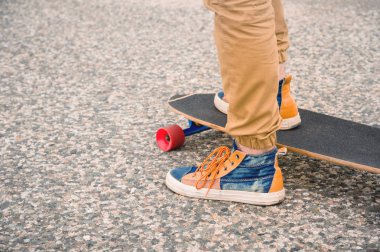 Güneş batımında asfalt arka planda bir Longboard üzerinde bez spor ayakkabıları erkek bacaklar yakın çekim. Erkek bacakları olan büyük bir kaykay. Gençlik eğlence konsepti