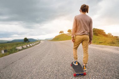 Uzun saçlı genç şık adam bir at kuyruğu ve güneş gözlüğü toplanan epik kayalar arka planda dağlarda bir ülke asfalt yol üzerinde bir Longboard ile duruyor