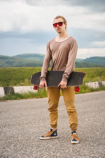 Ein stylischer junger Mann, der am Abend nach Sonnenuntergang mit Skate oder Longboard in der Hand an einer kurvenreichen Bergstraße steht. das Konzept des Jugendsports und der Reiselust — Stockfoto