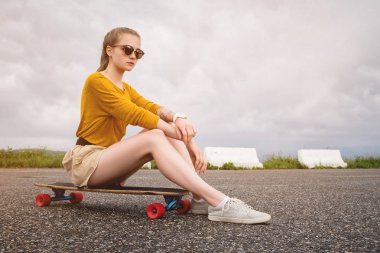 Güneş gözlüğü ve şort kolunda bir dövme ile Çekici genç kız gri gökyüzü bir arka plan üzerinde bir banliyö park onu longboard oturur. Millennials nesil ve eğlence kavramı