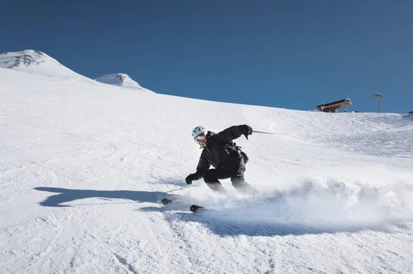 Un esquiador macho maduro barbudo con un traje de esquí negro desciende a lo largo de la ladera nevada de una estación de esquí en medio de dos picos del Monte Elbrus. El concepto de deporte en la edad adulta — Foto de Stock