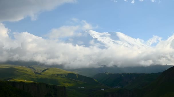 4K χρονοπαρέλθουν σύννεφα στροβιλίζονται σε ένα χιόνι καλυμμένο βουνό που κοιμάται ηφαίστειο Elbrus κατά το ηλιοβασίλεμα. Πράσινοι λόφοι με φαράγγια στο προσκήνιο. Βόρειος Καυκάσου Ρωσία. Αναρρίχηση στο βουνό από το Βορρά — Αρχείο Βίντεο