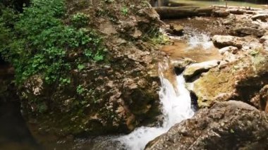 Hasır şapkalı genç bir adam ve çıplak gövdeli şort ve yalınayak bir sırt çantası yla bir orman dağ deresi ve büyük bir epik şelalenin yanında büyük bir kaya taşının üzerinde oturuyor. Havadan görünüm 4k seyahat videosu