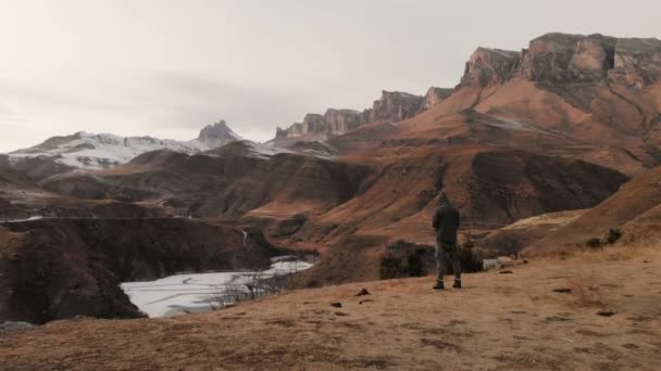 Een luchtfoto van een man in een kap staat aan de rand van een klif in de buurt van een bergmeer tegen de achtergrond van epische rotsen en bergen na zonsondergang in de schemering in de winter. — Stockvideo