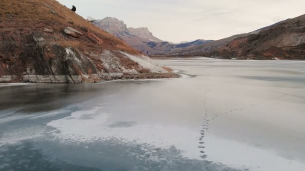 Vista aérea de una fotógrafa caminando a lo largo de la orilla de un lago de montaña congelado contra el telón de fondo de rocas épicas y montañas después del atardecer al atardecer en invierno . — Vídeo de stock