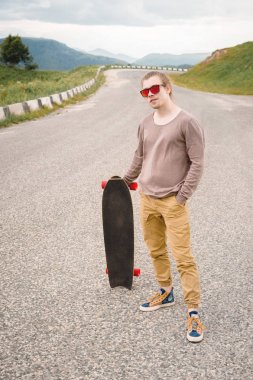 Şık bir genç adam gün batımından sonra akşam elinde bir paten veya longboard ile dolambaçlı bir dağ yolu boyunca ayakta. Gençlik spor ve seyahat hobileri kavramı