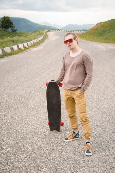 Ein stylischer junger Mann, der am Abend nach Sonnenuntergang mit Skate oder Longboard in der Hand an einer kurvenreichen Bergstraße steht. das Konzept des Jugendsports und der Reiselust — Stockfoto