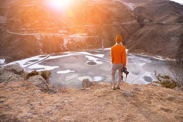 Porträtt från baksidan av flickan Traveler fotograf i en orange tröja och mössa med en kamera i handen i bergen mot bakgrund av en frusen Fjällsjö. Foto resekoncept — Stockfoto