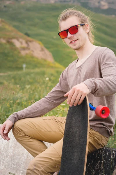 Homem elegante jovem com cabelos longos em óculos de sol está sentado em um chipper com um longboard em suas mãos em uma estrada de asfalto país no fundo de rochas e nuvens — Fotografia de Stock