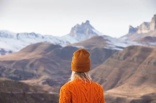 Retrato da parte de trás da menina viajante em uma camisola laranja e chapéu nas montanhas contra o fundo de uma montanha congelada. Foto conceito de viagem — Fotografia de Stock