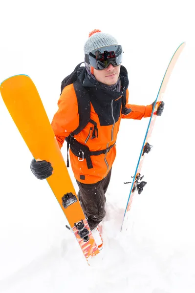 A professional skier athlete in an orange black suit with a black ski mask with skis in his hands stands with a displeased mass during a snowstorm on a light background in the snow