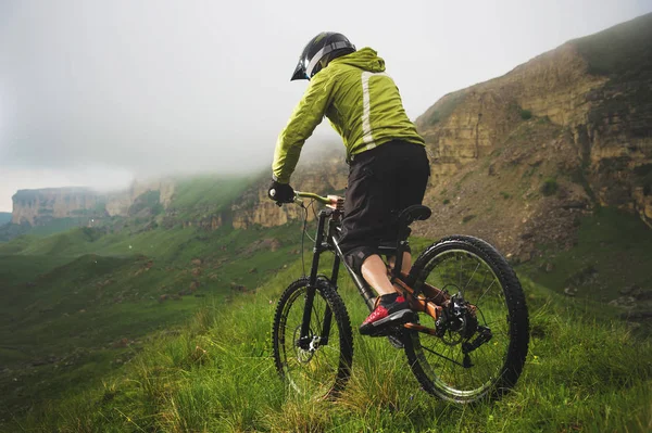 Hombre atleta envejecido en casco y máscara monta una bicicleta de montaña en una ladera cubierta de hierba contra el fondo de rocas meseta y nubes bajas en un día nublado. concepto de bicicleta de montaña cuesta abajo —  Fotos de Stock
