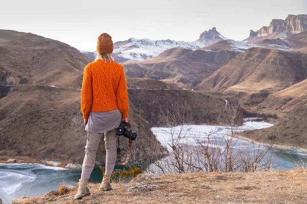 Portrait from the back of the girl traveler photographer in an orange sweater and hat with a camera in hand in the mountains against the background of a frozen mountain lake. Photo travel concept