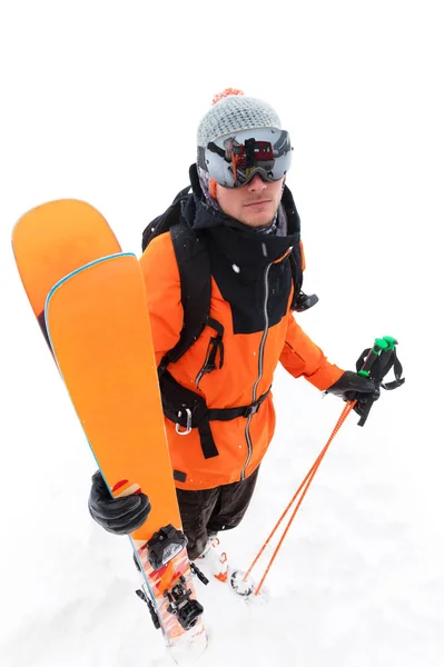 一名身着橙色黑色西装、手拿滑雪板、手拿滑雪板的专业滑雪运动员在暴风雪中站在雪地上,在浅色背景下站立着一个不满的群众。 — 图库照片