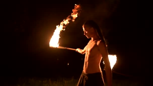 Zurückhaltend. junger Mann mit langen Haaren und nacktem Oberkörper dreht brennende Fackel im Freien auf einem schwarzen Nachtvideo in Zeitlupe. moderner Fakir trickst mit brennendem Stab — Stockvideo