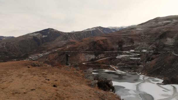Una vista aérea de un hombre en una capucha se encuentra en el borde de un acantilado cerca de un lago de montaña en el telón de fondo de rocas épicas y montañas después del atardecer al atardecer en invierno . — Vídeo de stock