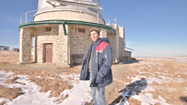 Um pesquisador do sexo masculino em jeans e um uniforme quente em um dia ensolarado de inverno contra o pano de fundo de edifícios científicos em um observatório solar. Estação de Pesquisa Científica nas Montanhas do Cáucaso — Vídeo de Stock