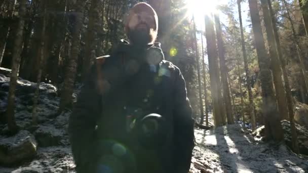 Portret van een ernstig bebaarde man fotograaf met een rugzak en een camera staande in een naald winter bos in achtergrondverlichting en zonlicht — Stockvideo