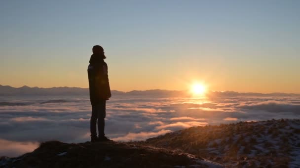 Η σιλουέτα ενός άντρα στέκεται στο ηλιοβασίλεμα πάνω από τα σύννεφα και σκέφτεται τον ήλιο που δύει. Η έννοια της ηρεμίας και της ευχαρίστησης στο ταξίδι — Αρχείο Βίντεο
