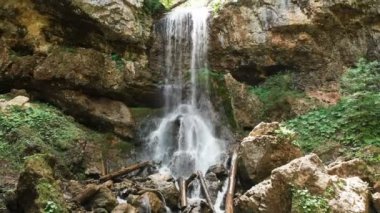 Hasır şapkalı genç bir adam ve çıplak gövdeli şort ve yalınayak bir sırt çantası yla bir orman dağ deresi ve büyük bir epik şelalenin yanında büyük bir kaya taşının üzerinde oturuyor. Havadan görünüm 4k seyahat videosu