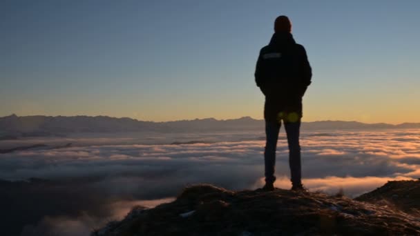 Vista pelas traseiras. Um homem de pé solitário no alto das montanhas olha para o pôr-do-sol e o horizonte do pôr-do-sol com um vale cheio de nuvens. O conceito de viagem turística e solidão masculina — Vídeo de Stock