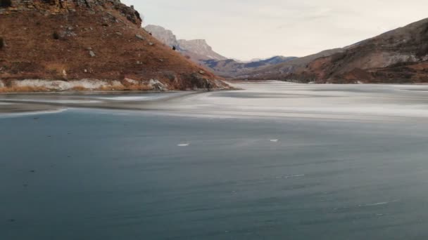 Vista aérea de una fotógrafa caminando a lo largo de la orilla de un lago de montaña congelado contra el telón de fondo de rocas épicas y montañas después del atardecer al atardecer en invierno . — Vídeo de stock