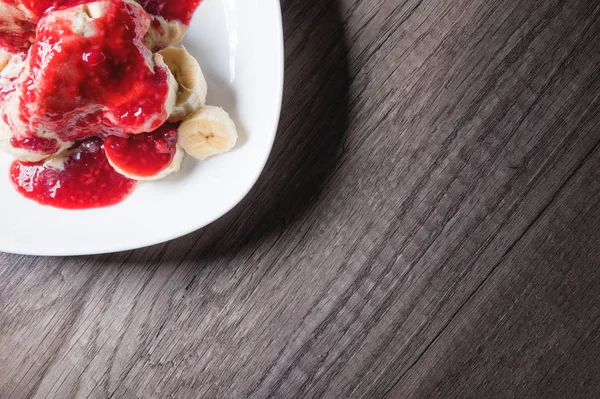 Closeup Vanilla ice cream poured with strawberry or raspberry jam with sliced banana slices on a white plate on a dark wooden table. You can do this at home on vacation