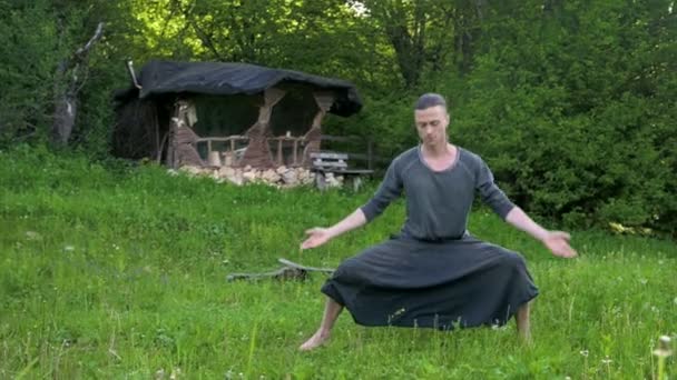 Algemeen plan een jonge man in Indiase kleding en broek Aladdin praktijken Qigong terwijl het doen van oefeningen werken met Qi energie in de zomer buiten naast een oude hut 4k — Stockvideo