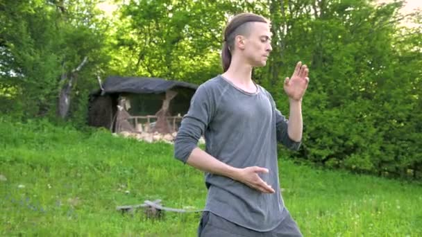 Un plan moyen un jeune homme en vêtements et pantalons indiens Aladdin pratique le qigong tout en faisant des exercices tout en travaillant avec l'énergie qi à l'extérieur en été à côté d'une vieille cabane — Video