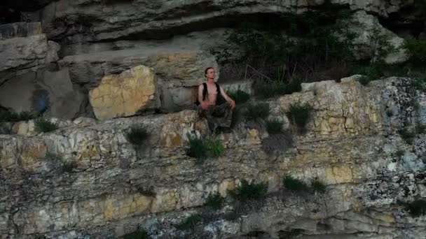 Aerial view of a traveler with a backpack sitting on the edge of a shelf in the rock and looking into the distance. 4K epic travel video — Stock Video