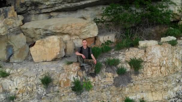 Vista aérea de un viajero con una mochila sentada en el borde de un estante en la roca y mirando a la distancia. Vídeo épico de viaje 4K — Vídeo de stock