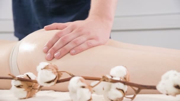 Närbild av en manlig sjukgymnast gnugga massage olja på låret och skinkorna på en ung kvinna innan massagebehandlingar i en professionell Spa salong. Koncept för avslappning och kroppsvård i 4K — Stockvideo