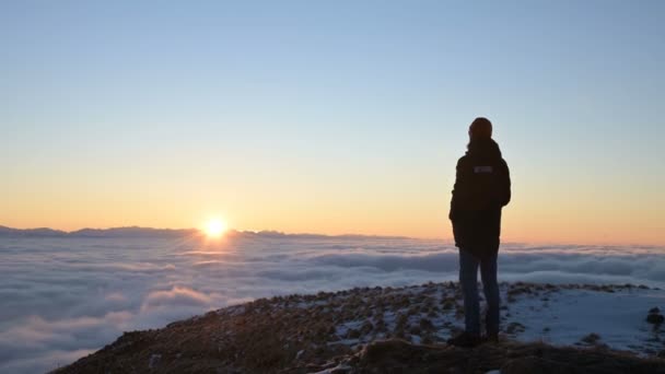 Vista desde atrás. Un solitario hombre de pie en lo alto de las montañas mira el sol poniente y el horizonte del atardecer con un valle lleno de nubes. El concepto de viajes turísticos y la soledad masculina — Vídeo de stock