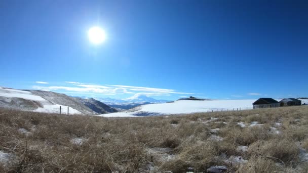 Timelapse dag flera hus mot bakgrund av snötäckta berg i Kaukasus med bildandet av moln i den klara himlen. Solens rörelse på himlen — Stockvideo