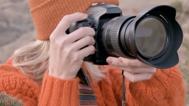 Close-up fotógrafo viajante menina em um suéter laranja e chapéu tira fotos em sua paisagem câmera dslr no outdor contra o pano de fundo de montanhas épicas. Viagens Fotógrafos conceito de vídeo — Vídeo de Stock