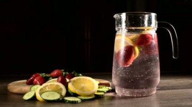 Taze meyve ve köpüklü maden suyu limon nane ve salatalık ile dondurulmuş çilek ile karanlık bir arka plan üzerinde Cam karafe. Köpüklü suya meyve atmak