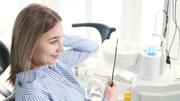 Retrato de una chica sonriente rubia en una silla dental con un espejo en forma de diente en sus manos. Gabinete dental cliente feliz — Vídeos de Stock