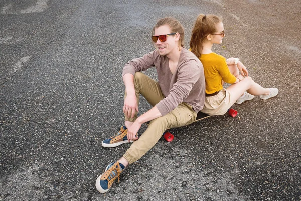 Ein attraktives junges Paar mit Sonnenbrille lehnt sich Rücken an Rücken auf seinem Longboard auf einem Vorstadtparkplatz vor grauem Himmel zurück. das Konzept einer jungen Generation Millennials Stil und Freizeit — Stockfoto