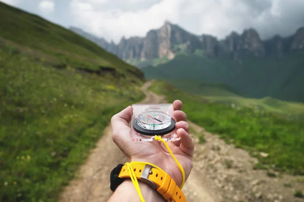 Zbliżenie szeroki kąt męski ręka z żółtym zegarku zegarek posiada magnetyczny kompas nawigacji na tle pięknego krajobrazu w górach z brudną drogą krowa. — Zdjęcie stockowe