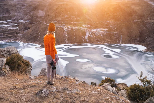 Porträtt från baksidan av flickan Traveler fotograf i en orange tröja och mössa med en kamera i handen i bergen mot bakgrund av en frusen Fjällsjö. Foto resekoncept — Stockfoto