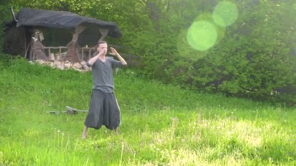 General plan en ung man i indiska kläder och byxor Aladdin Practices Qigong medan du gör övningar arbeta med Qi energi på sommaren utomhus bredvid en gammal Hut 4K — Stockvideo