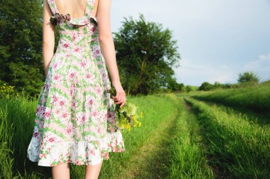 Kır çiçekleri bir buket ile hafif bir yaz elbise li bir kızın arkasından bir görünüm bir kır yolu önünde duruyor. Kadınların eşit haklar ve bağımsızlığı feminizm kavramı