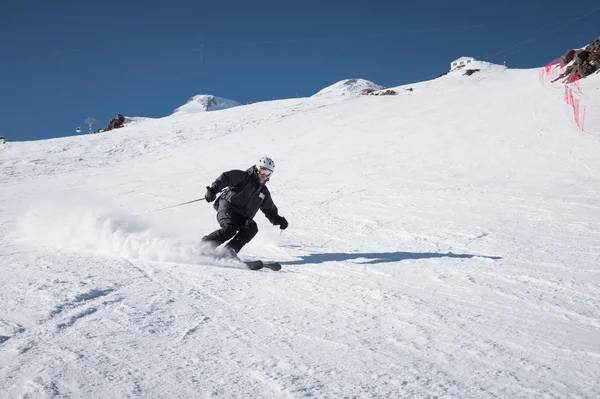 Un esquiador macho maduro barbudo con un traje de esquí negro desciende a lo largo de la ladera nevada de una estación de esquí en medio de dos picos del Monte Elbrus. El concepto de deporte en la edad adulta — Foto de Stock