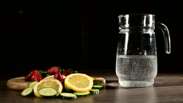 Glazen karaf op een donkere achtergrond met vers fruit en bevroren aardbeien met citroen munt en komkommers in bruisend mineraalwater. Giet bruisend water in een glazen karaf — Stockvideo